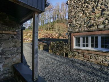 A gravel driveway between stone buildings with a window on one building and trees in the background at Church How Cottage in Nether Wasdale