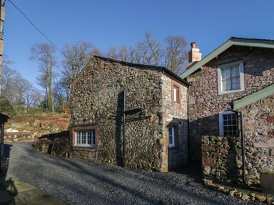 A stone cottage with multiple windows and a gravel driveway surrounded by leafless trees at Church How Cottage in Nether Wasdale