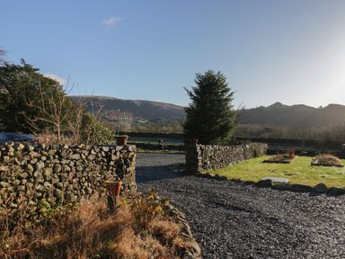 A gravel driveway entrance with stone walls and garden beds with mountains in the background at Church How Cottage in Nether Wasdale