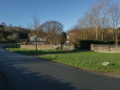 A country road next to a grassy area with a stone wall bench and trees at Church How Cottage in Nether Wasdale