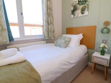 A bedroom with a single bed with white bedding and green leaf patterned cushion and curtains a bedside table with flowers and a lamp at Daffodil Lodge Mullacott near Ilfracombe