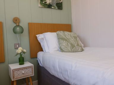 A bedroom with a bed with white sheets and a green patterned pillow next to a small bedside table with a green vase and flowers at Daffodil Lodge in Mullacott near Ilfracombe