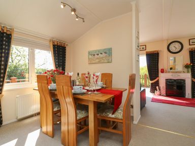 A dining area with a wooden table set for six next to a window and a living room with a fireplace and clock at Poppy Lodge in Mullacott Ilfracombe