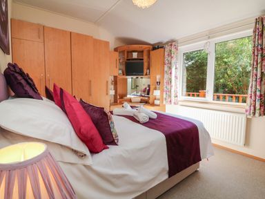 A bedroom with a bed with purple pillows and runner wooden wardrobes and a window with floral curtains at Poppy Lodge Mullacott Ilfracombe