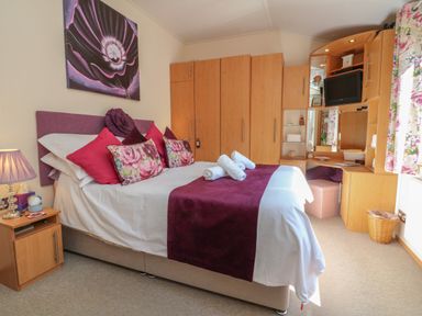 A bedroom with a double bed with purple and floral pillows and bedding wooden wardrobes and a vanity unit at Poppy Lodge Mullacott Ilfracombe