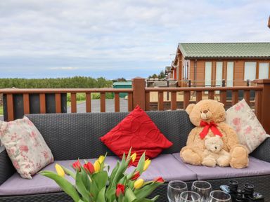 An outdoor seating area with a wicker couch, floral and red cushions, two teddy bears, tulips on a table, glassware, and a green-roofed cabin in the background at Poppy Lodge Mullacott Ilfracombe