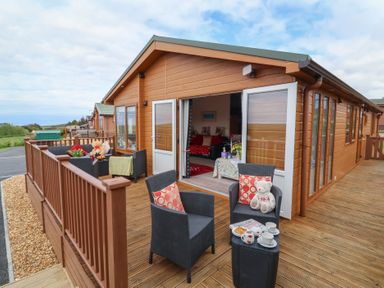 An outdoor wooden deck with black wicker chairs and a small table in front of a wooden lodge with open doors at Poppy Lodge in Mullacott Ilfracombe