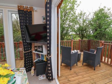 A small living area with a wall-mounted television and shelves beside a deck with two wicker chairs and a small table at Poppy Lodge in Mullacott Ilfracombe
