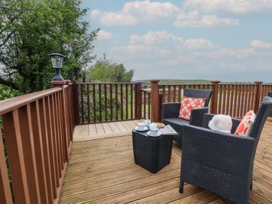 A wooden deck with two black wicker chairs and a small table set with cups and a teapot at Poppy Lodge in Mullacott Ilfracombe