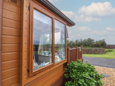 A wooden cabin exterior with a large window showing indoor decorative items and a garden area outside at Poppy Lodge in Mullacott Ilfracombe