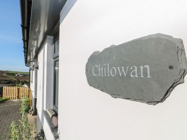 A close up of a stone house name plaque on a white wall with a window and outdoor area at Chilowan in Trewarmett near Tintagel