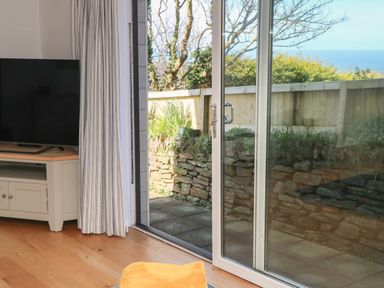 A living room with a television on a stand and sliding glass doors opening to a patio with a stone wall at Chilowan in Trewarmett near Tintagel