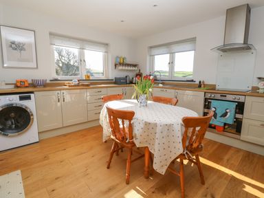 A kitchen with wooden chairs around a table with a tablecloth and a vase of flowers with cabinets along the walls and a washing machine Chilowan Trewarmett near Tintagel