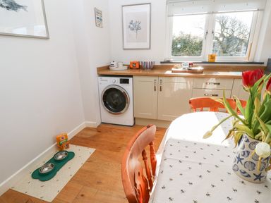A kitchen corner with a washing machine under a wooden countertop and a dining table with a vase of flowers at Chilowan in Trewarmett near Tintagel