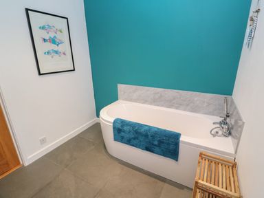 A bathroom with a bathtub with a blue towel a wooden stool a framed fish picture on the wall and teal and white walls at Chilowan in Trewarmett near Tintagel