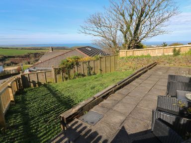A garden with a paved patio and wicker chairs overlooking houses fields and the sea at Chilowan Trewarmett near Tintagel