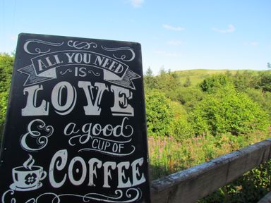 A sign with a coffee quote outdoors with green trees and hills in the background at Cuckoo at Wild Valley Huts in Llanrhaeadr-Ym-Mochnant
