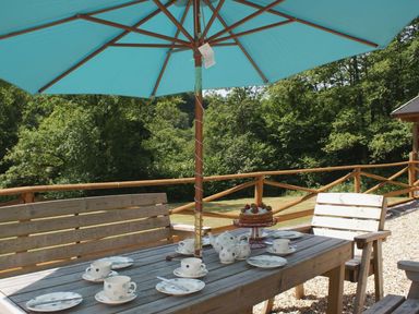 An outdoor wooden table set with cups plates teapot and cake under a blue umbrella surrounded by wooden chairs on a deck at Valley Lodge Oakfordbridge near Bampton