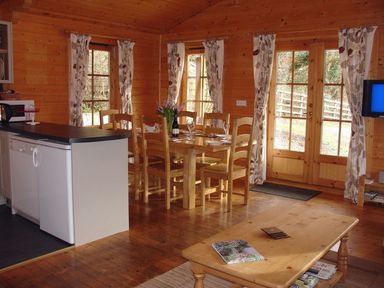 A dining area with wooden chairs and table set with glasses and a bottle next to kitchen counter and coffee table in a wooden room at Valley Lodge Oakfordbridge near Bampton