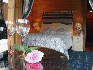 A bedroom with a bed and patterned bedding two bedside tables with lamps and flowers and a table with wine glasses rose and wine bottle at Valley Lodge in Oakfordbridge near Bampton