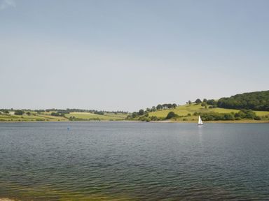 A lake with a sailboat and hills with trees and fields in the background at Valley Lodge in Oakfordbridge near Bampton