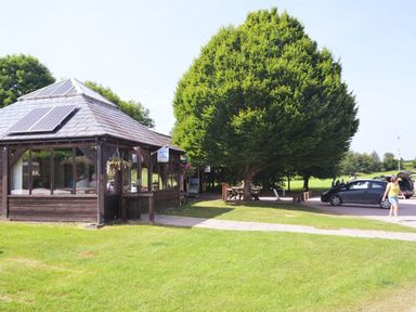 A wooden pavilion with solar panels and large trees beside a parking area with cars and people at Valley Lodge in Oakfordbridge near Bampton