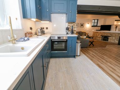 A kitchen with blue cabinets a white countertop a brass faucet and appliances next to a living room with a sofa a TV and a stone fireplace at Ellie's Cottage in Burford