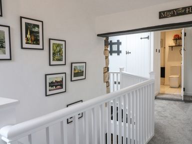 A staircase with white railing and framed pictures on the wall leading to a hallway with a bathroom in Ellie's Cottage Burford