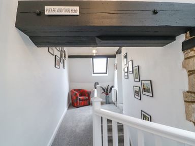 A stairway landing with a red armchair a small table a vase with plants framed pictures on white walls and a low black wooden beam at Ellie's Cottage in Burford