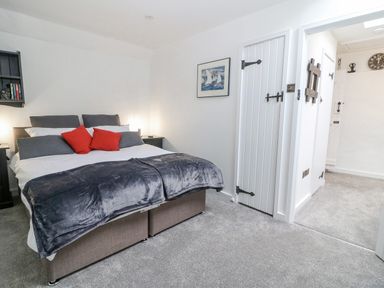 A bedroom with a bed with grey and red pillows and a grey blanket with white walls and carpet at Ellie's Cottage in Burford