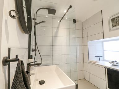 A bathroom with a sink a black faucet a round mirror a shower area and a window with toiletries at Ellie's Cottage in Burford