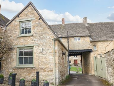 A stone house with green framed windows and an archway leading to a street at Ellie's Cottage in Burford