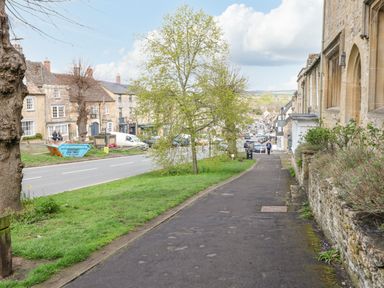 A paved sidewalk beside a stone building with plants and trees along a street with parked cars and houses in Burford