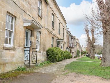 A row of stone cottages along a cobblestone path with trimmed bushes and leafless trees on a grassy verge at Ellie's Cottage in Burford