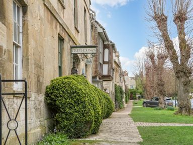 A row of stone buildings with a greyhounds b and b sign and manicured bushes along a cobbled sidewalk with parked cars and leafless trees nearby at Ellie's Cottage in Burford