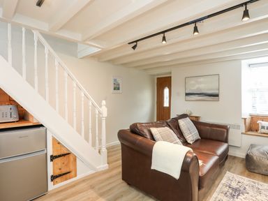 A living room with a brown leather sofa pillows and a white blanket near a wooden door and white staircase at Bwthyn Lili (Lily Cottage) in Cemaes Bay