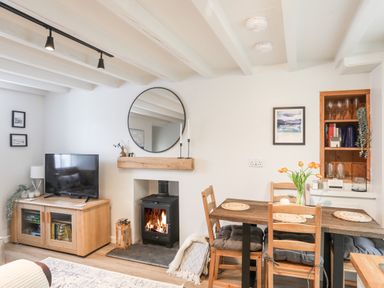 A living room with a wood stove next to a dining table with four chairs and a TV on a wooden cabinet at Bwthyn Lili (Lily Cottage) in Cemaes Bay