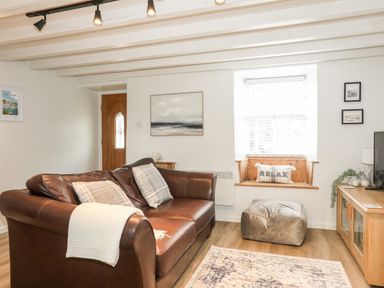 A living room with a brown leather sofa two plaid cushions a white throw a window seat with a relax cushion a beige pouf a wooden media cabinet and a rug at Bwthyn Lili Cemaes Bay