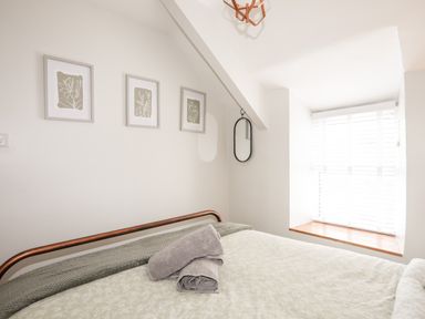 A bedroom with a bed, towels on the bed, three framed pictures on the wall, an oval mirror, and a window with blinds at Bwthyn Lili (Lily Cottage) in Cemaes Bay