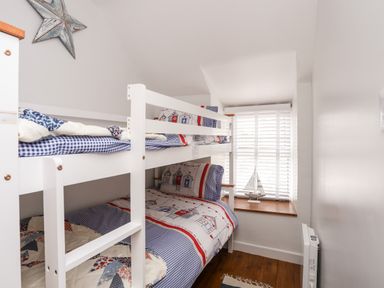 A small bedroom with white bunk beds bedding with house patterns a window with blinds and a model sailboat on the windowsill at Bwthyn Lili (Lily Cottage) in Cemaes Bay