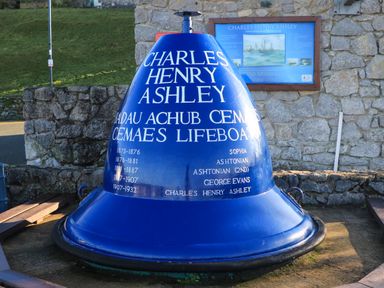 A large blue lifeboat bell painted with white text about Charles Henry Ashley placed outdoors near a stone wall at Bwthyn Lili (Lily Cottage) in Cemaes Bay