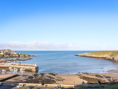 A coastal harbor with boats docked near a pier and a beach with rocky areas and people walking at Bwthyn Lili (Lily Cottage) in Cemaes Bay