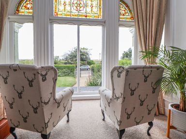Two armchairs with deer head patterns in front of a large window with stained glass panels and curtains in a sitting room with a plant in a pot at Ismay Dining Room Suite - Titanic Interest in Waterloo Merseyside