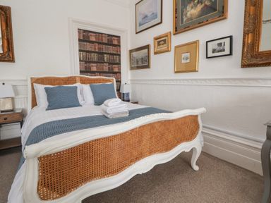 A bedroom with a bed with woven headboard and footboard two blue cushions and a stack of towels on the bed nightstands with lamps and framed pictures on the walls at Ismay Dining Room Suite - Titanic Interest in Waterloo Merseyside