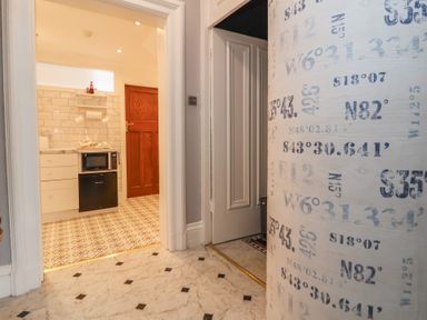 A hallway with marble flooring and a curved wall with coordinates leading to a kitchen with patterned tile floor and wooden door at Ismay Dining Room Suite - Titanic Interest in Waterloo Merseyside