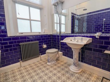 A bathroom with blue tiled walls a patterned floor a toilet a pedestal sink a radiator and a window at Ismay Dining Room Suite - Titanic Interest in Waterloo Merseyside