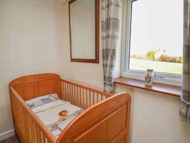 A baby crib with animal print bedding next to a window with curtains and a vase on the windowsill at Brynteg Cottage in Tegryn