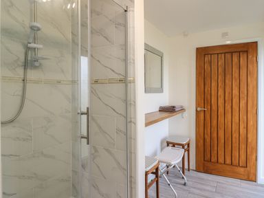 A shower area with tiled walls and a glass door next to a wooden door with two stools under a wooden shelf with folded towels and a mirror on the wall at Brynteg Cottage in Tegryn
