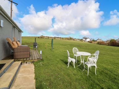 A patio with wicker chairs and cushions next to a grassy lawn with a white metal table and four chairs at Brynteg Cottage in Tegryn