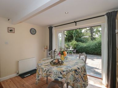 A dining area with a round table covered with a patterned tablecloth and flowers by a large open window with a view of an outdoor table and chairs at Nuthatch in Honiton